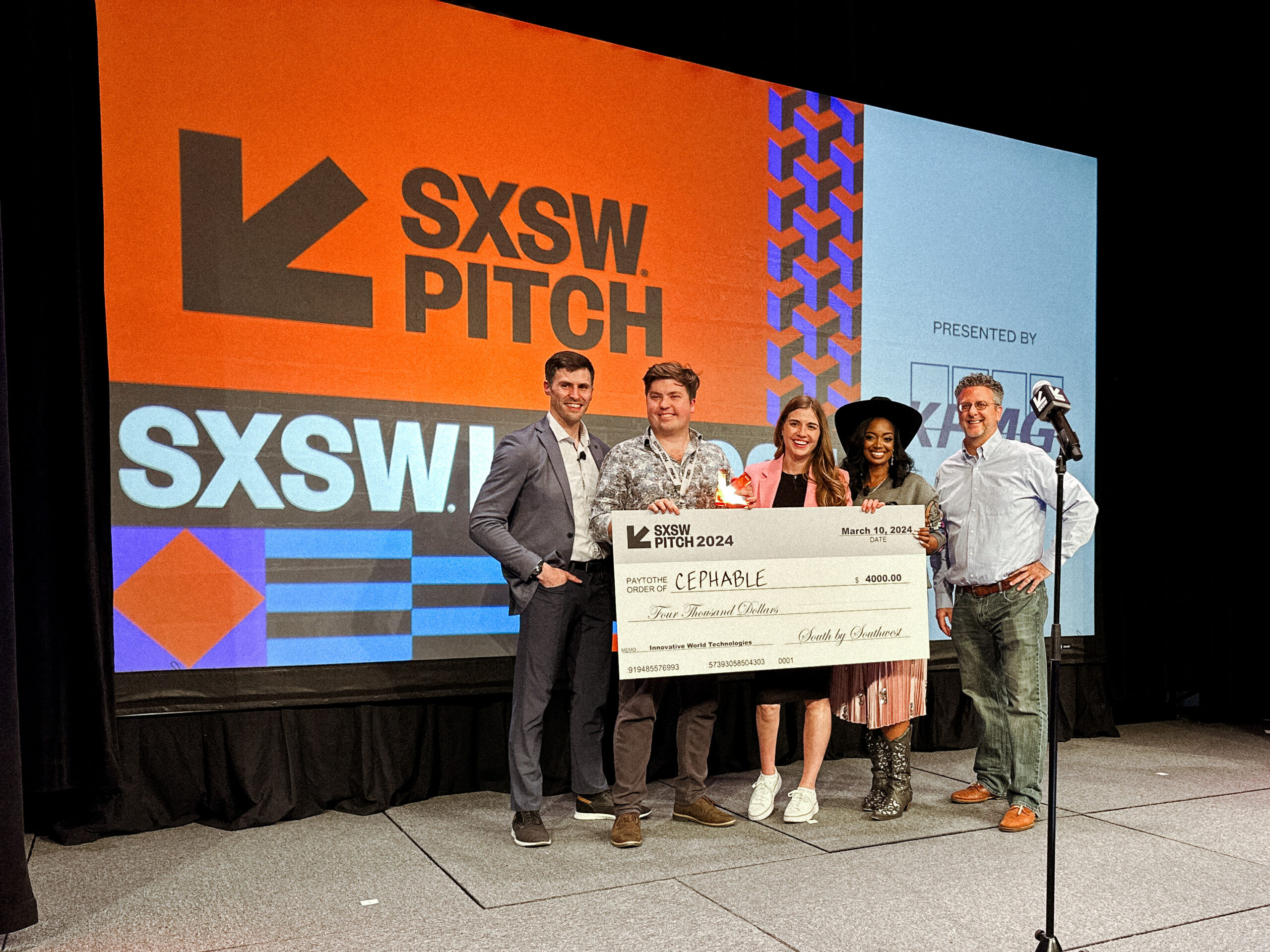 Cephable CEO Alex Dunn and Director of Marketing Alexa Orban stand on the SXSW Pitch stage holding an award and check. There are 3 members of the SXSW team on stage in alongside them and an orange SXSW Pitch banner in the background. 