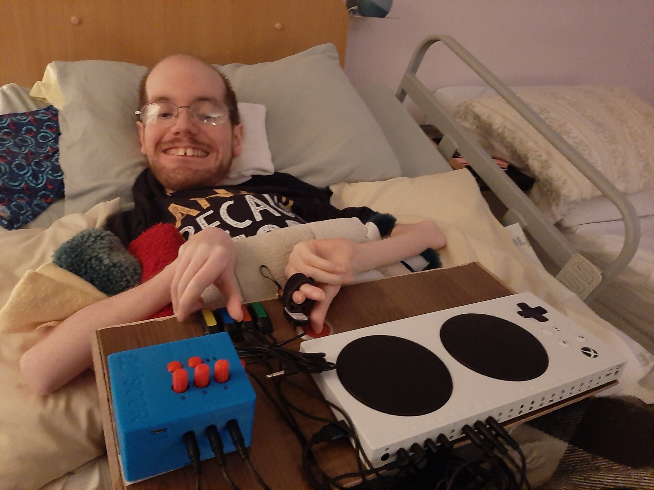 A disabled gamer is lying in bed, smiling, and using an adaptive gaming setup. The setup includes a custom blue control box with large red buttons and a white adaptive controller with two large black buttons. 