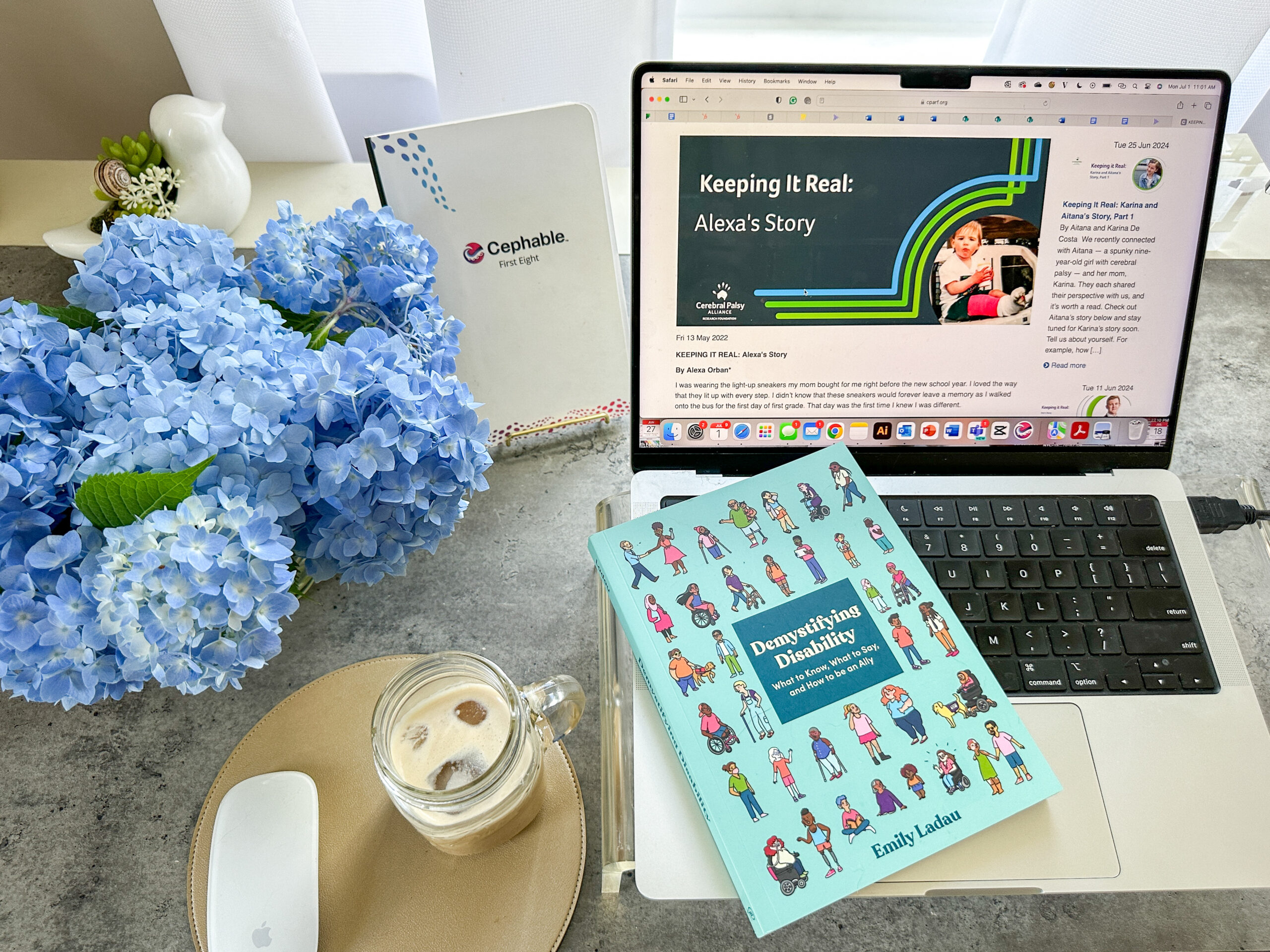 A photo of Alexa's desk. On the left, a bouquet of blue hydrangeas sits next to a cup of iced coffee, a mouse, and a Cephable journal. Alexa's laptop is on the right, with a screen that shows 'Keeping it Real: Alexa's Story' from the Cerebral Palsy Alliance Research Foundation. Demystifying Disability sits on the keyboard.