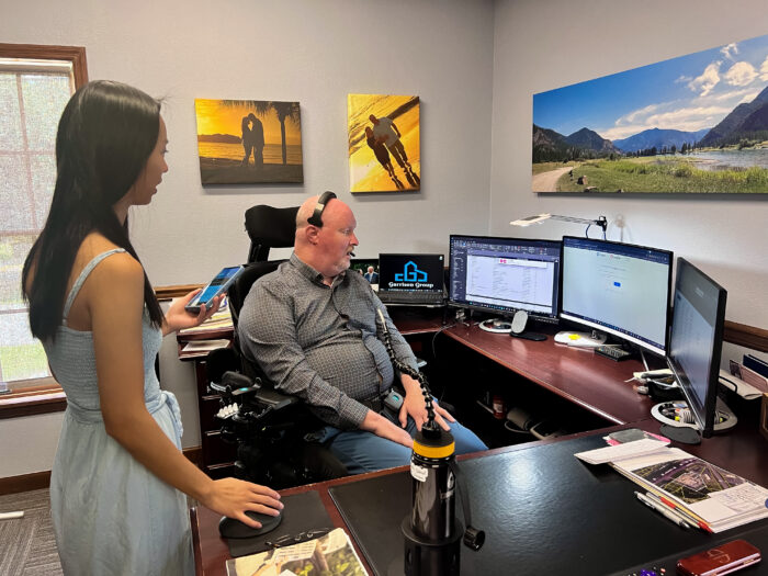 An image of Alexa and Rex in his office. Rex is using a power chair and sitting at his computer that has 3 monitors. 