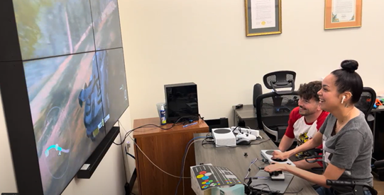 Zach sits with a player who is using an accessible controller while playing a racing video game. 