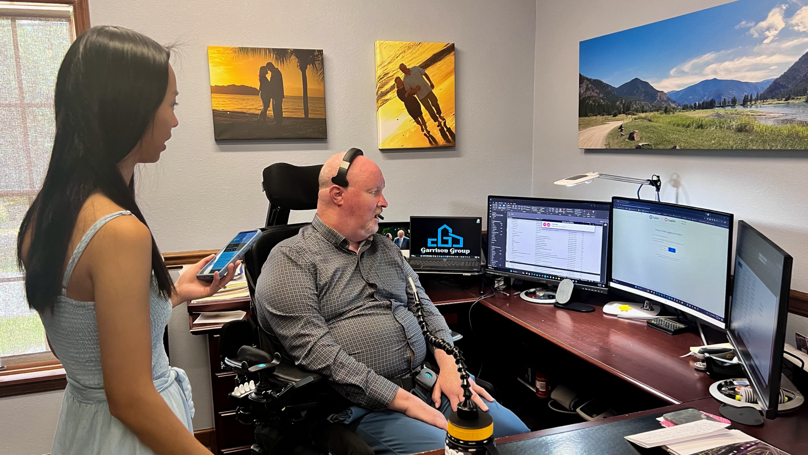 An image of Alexa and Rex in his office. Rex is using a power chair and sitting at his computer that has 3 monitors.